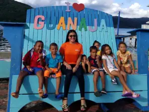 Scenic view of Dunbar Rock with a group of children and a woman sitting on a large blue boat-shaped structure with colorful letters spelling 'GUANJA' in the background.